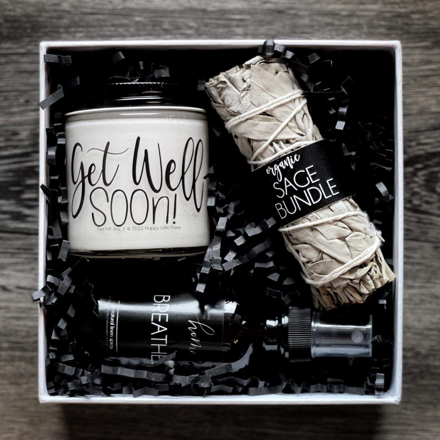 Open box of the pre-wrapped get well soon kit from Happy Little Flame which includes a white sage smudge stick , a lavender vanilla candle, and an all-natural breathe spray.